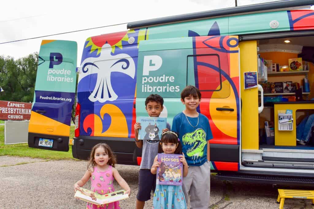 Dos niñas y dos niños sonriendo con libros ilustrados delante de una colorida furgoneta.