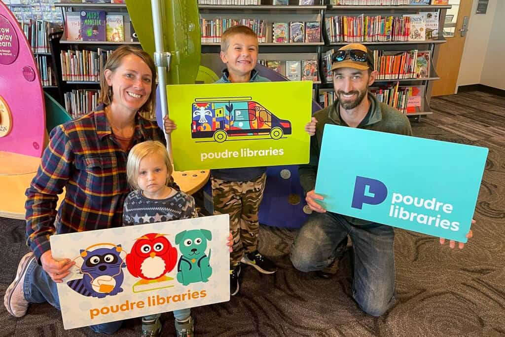 Madre y padre sonriendo con sus dos hijos pequeños sosteniendo coloridos carnés de biblioteca de gran tamaño.