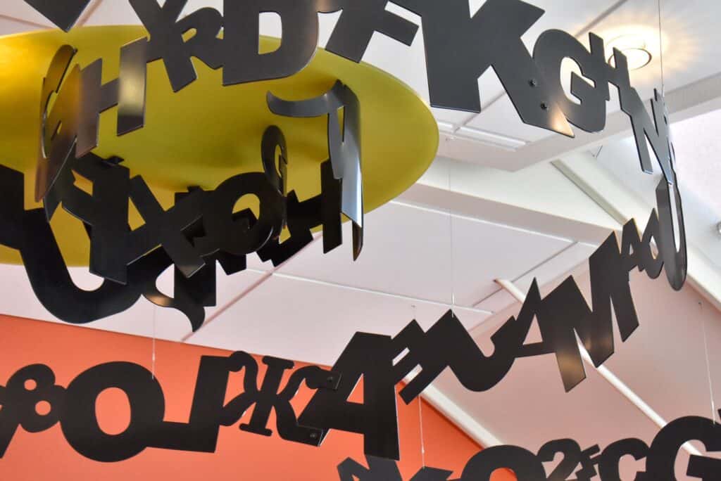 Large hanging black metal sculpture with different letters circling a yellow base