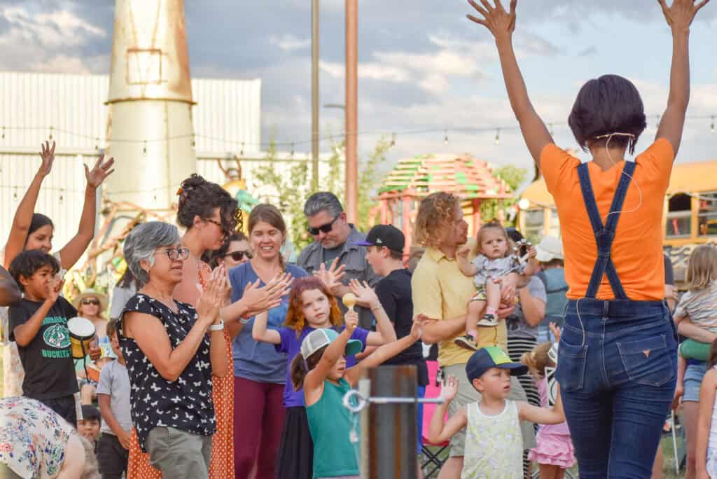 The back of a performer with her hands up facing a diverse group of adults and children at an outdoor event.