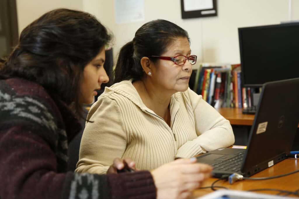 Mujer gafas mirando un portátil junto a una mujer más joven que la asiste