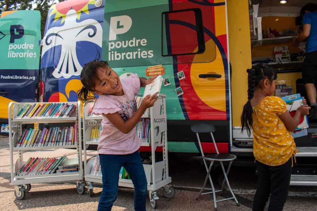 Young girl jumping up with a book in her hands outside a colorful library bookmobile