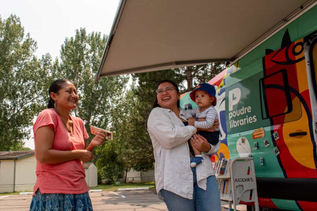 Two women, one holding a baby, laughing in front of a colorful library bookmobile