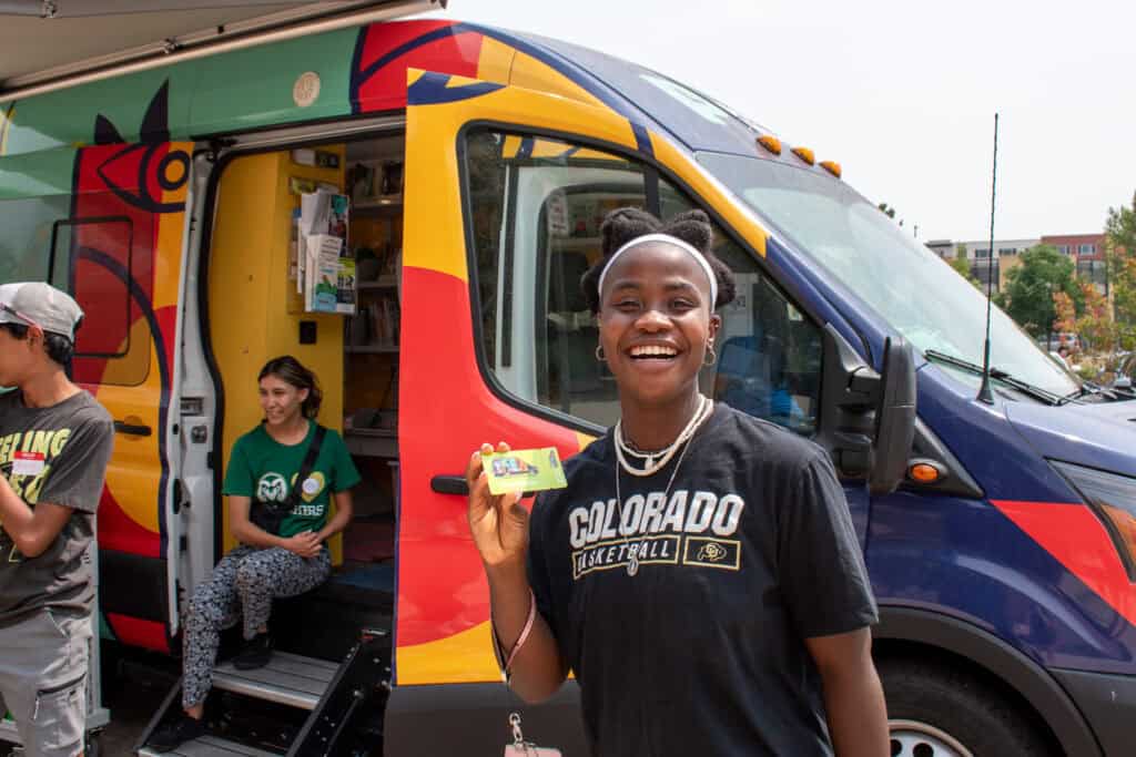 Young woman smiling with a new library card outside a colorful library bookmobile