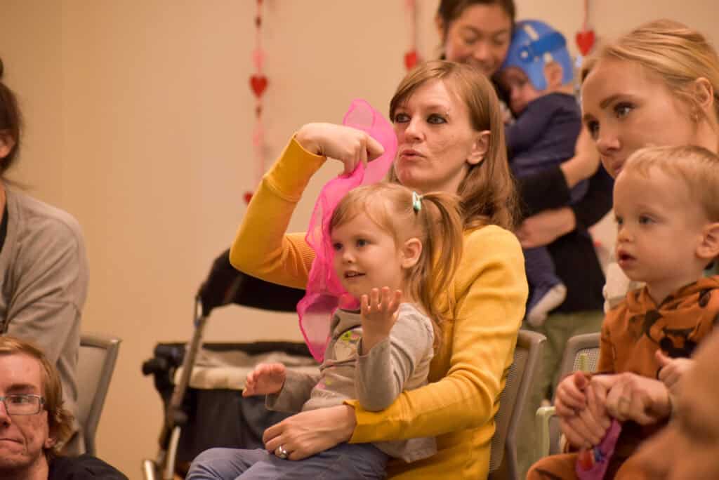 Madre con su hija en el regazo jugando con un pañuelo rosa en la hora del cuento de la biblioteca