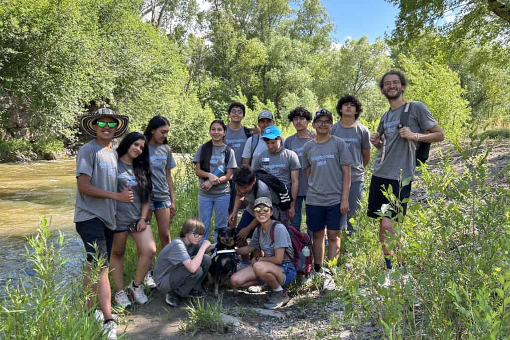 A group of teens and an adult man smiling for a photo by a river