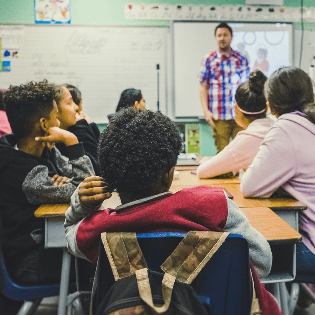 Back of a young boys head sitting in a classroom with his peers and looking at his teacher