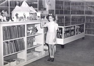 Black and white image of a little girl standing by a shelf of books in a library