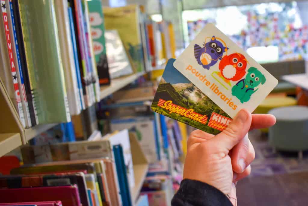 Hand holding up a poudre libraries card and a king sooper's shopper card together