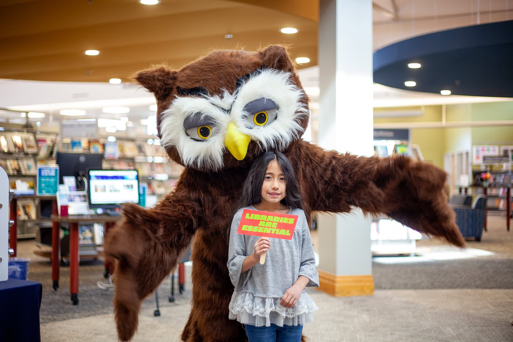 girl holding sign that reads libraries are essential while posing for a photo with an owl mascot
