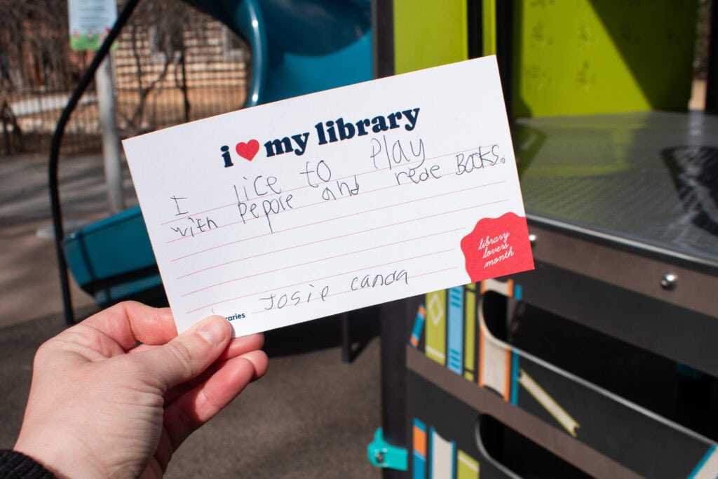 hand holding up a notecard with a child's writing that says "i lice to play with people and rede books"