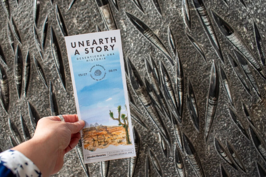 hand holding up a summer adventure bookmark in front of a rock with fossils in it