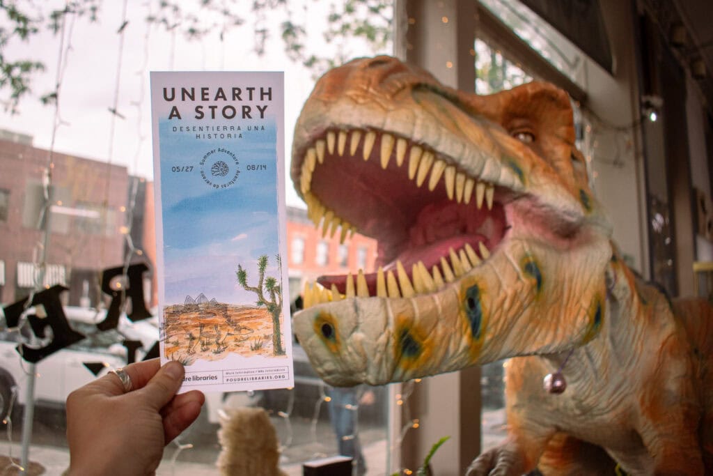 mano mostrando un marcapáginas de aventura de verano delante de un dinosaurio